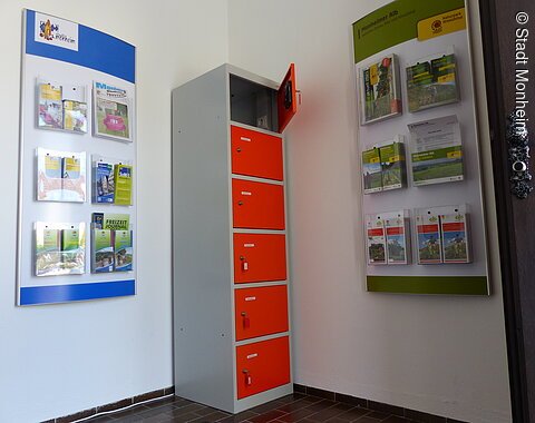 Ein orange-grauer Indoor-Ladeschrank für eBike-Akkus mit insgesamt sechs Fächern und einem offenen Fach steht zwischen zwei Wanddisplays mit Broschüren in dem Vorraum der Touristinformation Monheim.