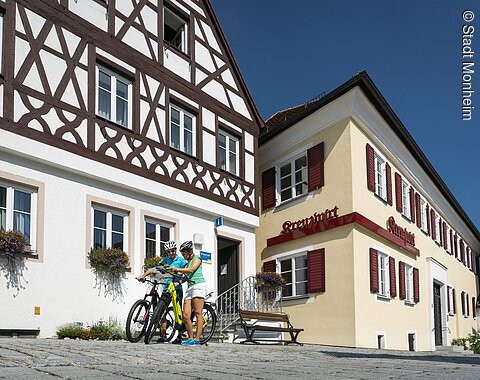 Tourist-Information der Stadt Monheim/Monheimer Alb: ein Fachwerkhaus mit drei Stockwerken, weißen Wänden und braunen Balken. Rechts davon ein gelbes zweistöckiges Gebäude mit roten Fensterläden. Zwei Personen mit Fahrrädern stehen davor und schauen eine Broschüre an.