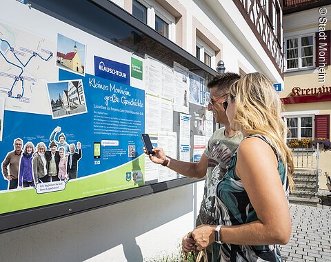 Zwei Personen vor einem weißen Gebäude, der Touristinformation Monheim betrachten eine Informationstafel mit einer Karte und Texten zur Lauschtour in Monheim und haben ein Smartphone in der Hand, mit dem sie einen Code abscannen.