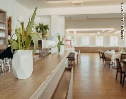 Weißer Blumenstrauß in Vase auf Holztheke in hellem, leerem Restaurant mit Tischen und Stühlen im Hintergrund.