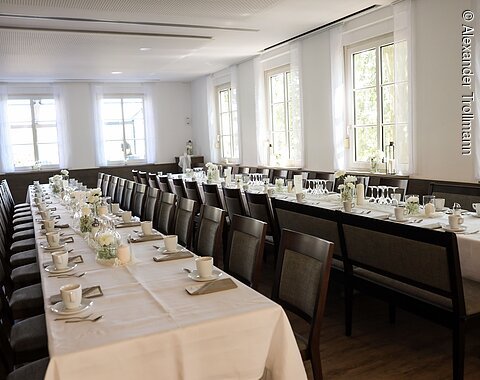 Langer gedeckter Tisch mit weißen Tassen, Gläsern und Blumen in einem hellen Raum mit Fenstern.