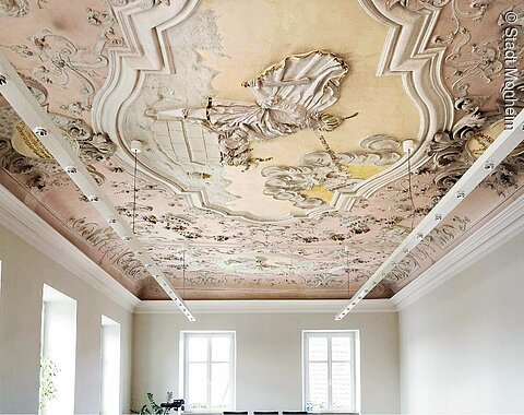 Farbig gestaltete Decke mit barocker Stuck- und Freskoverzierung in einem hellen Raum mit weißen Wände, vielen Fenstern und moderner an der Decke hängender Beleuchtung.