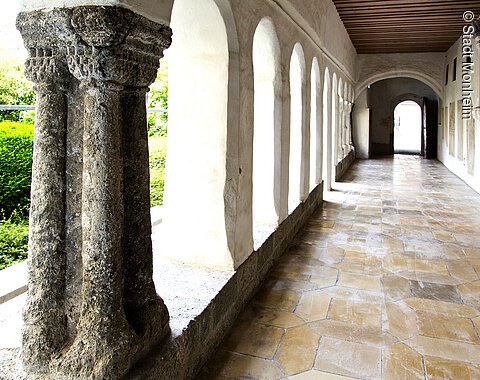 Blick in einen langen Kreuzgang mit Säulen und Rundbögen links im Bild, dahinter grüne Büsche. Rechts im Bild weiße Wand mit einer Ausfräsung in der Wand und Steinplatten mit Text angebracht. Entlang des Gangs alter Bodenbelag aus hellen alten Steinen.