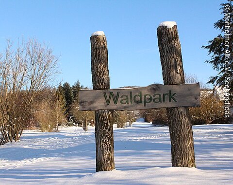 Willkommen im Waldpark Huisheim Holzschild mit der Aufschrift „Waldpark“ zwischen zwei Baumstämmen im verschneiten Park unter blauem Himmel
