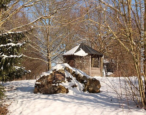 Waldpark Huisheim - Gedenkstein mit Infohäuschen Schneebedeckter Wald mit kahlen Bäumen, einem großen Stein mit Schild und einer Holzhütte im Hintergrund.