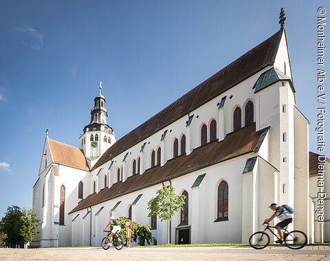 Blick auf das Kaisheimer Marienmünster Weißes Kirchgebäude mit Turm und zwei Radfahrern im Vordergrund bei blauem Himmel.