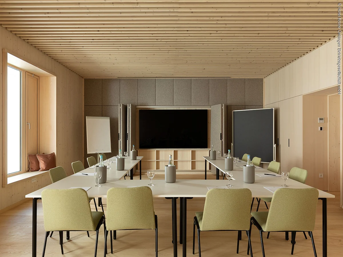 Moderner Konferenzraum mit U-förmigem Tisch, grünen Stühlen, Flipchart und großem Bildschirm an der Wand.