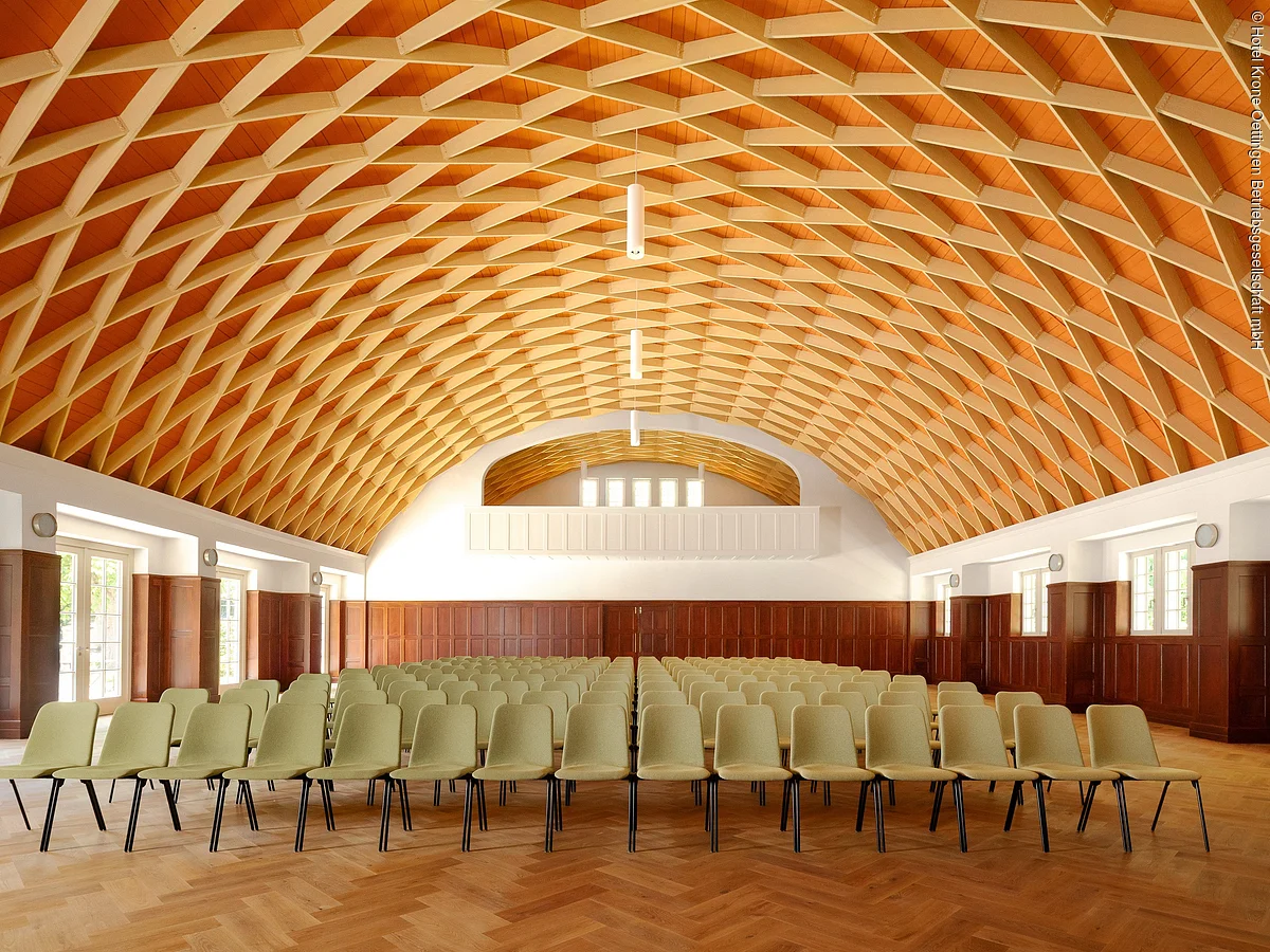 Großer Saal mit gewölbter Holzdecke und mehreren Reihen grüner Stühle auf Parkettboden.