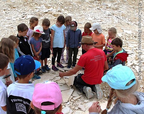 B+ Zentrum Blossenau - Besuch im Steinbruch Kinder stehen im Kreis um eine Person mit rotem Shirt und Hut, die auf dem Boden sitzend Steine bearbeitet.