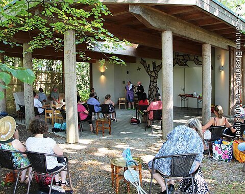 B+ Zentrum Blossenau - Musik erleben Menschen sitzen auf Stühlen im Freien unter einem Holzpavillon und hören zwei Personen zu, die vorne stehen.