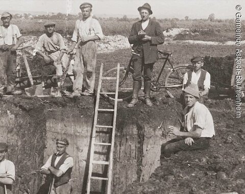 Neun Männer in Arbeitskleidung bei einer Ausgrabung mit Leiter und Schubkarre auf offenem Feld.