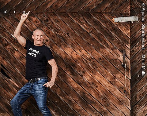 Michl Müller "Limbo of Life" in der Stadthalle Monheim Mann in schwarzem T-Shirt mit Aufschrift „DRIGG-SAGG“ und dunkelblauer Jeans posiert in die Kamera blickend vor Holztür mit diagonalem Muster.
