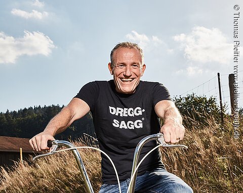 Michl Müller "Limbo of Life" Mann mit schwarzem T-Shirt mit der weißen Aufschrift "DREGG-SAGG" und Jeans sitzt lachend auf orangefarbenem Fahrrad auf Feldweg bei Sonnenschein
