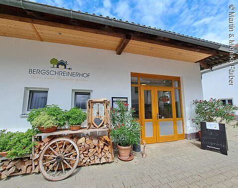 Eingang zum Gastronomiebereich des Bergschreinerhofs in Wolferstadt Eingangsbereich eines Hauses mit Holzdeko, Pflanzen und Schriftzug „Bergschreinerhof. Natur erleben. Regionales genießen.“ an der Wand.