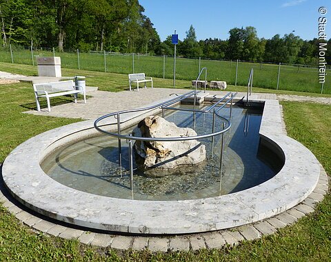 Rundes Kneippbecken in Monheim mit einem Geländer und einem Barfußpfad, in der Mitte ein großer weißer Stein. Umgeben von einer Wiese, darauf Sitzmöglichkeiten und eine große Grünfläche mit Bäumen hinterhalb, abgetrennt mit einem Zaun.