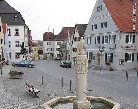 Blick zum Unteren Torturm vom Monheimer Markplatz Im Vordergrund ein grauer achteckiger Brunnen aus Jura-Stein mit einer Säule und einer darauf Sitzenden Skulptur des Adlers. Brunnen ist mit Wasser befüllt auf einem gepflastertem Platz mit Häusern und parkenden Autos am Straßenrand. Blumen links unten im Bild im Vordergrund.