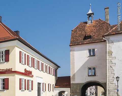 Tourist-Information der Stadt Monheim/Monheimer Alb - im Schindlerhaus Tourist-Information der Stadt Monheim/Monheimer Alb: ein Fachwerkhaus mit drei Stockwerken, weißen Wänden und braunen Balken. Davor steht ein Schaukasten und zwei Radfahrer fahren auf gepflasterter Straße vorbei. Rechts davon ein gelbes zweistöckiges Gebäude mit roten Fensterläden.