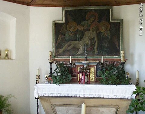 Innenansicht einer Kapelle: Altar mit weißem Tuch bedeckt, umgeben von verschiedenen Kerzen, Blumen und religiösem Gemälde oberhalb an der Wand, in einem Raum mit Holzbalkendecke.