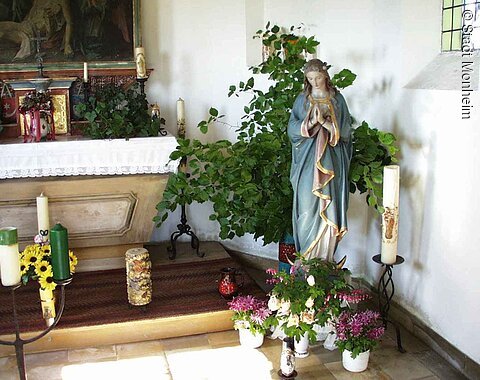 Innenansicht einer kleinen Kapelle mit der Statue einer betenden Frau mit blauem Mantel in einer Ecke umgeben von Kerzen, Pflanzen und Blumen in einem weißen Raum. Links im Bild ein geschmückter Altar.