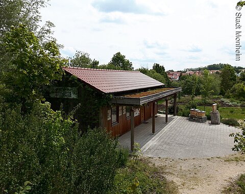 Holzgebäude mit rotem Dach und begrüntem Vordach, umgeben von Bäumen und einem gepflasterten Vorplatz, bei bewölktem Himmel.