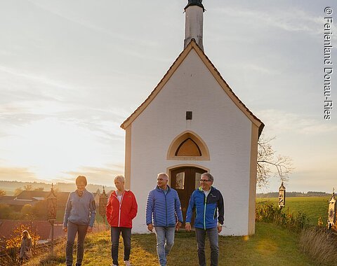 Vier Personen gehen nebeneinander auf einer Wiese vor einer kleinen Kapelle im Sonnenuntergang spazieren. Im Hintergrund grüne Wiese und weitere Gebäude.
