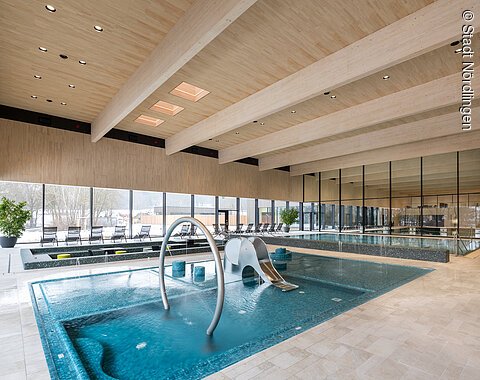 Blick auf das Kinderbecken im Kraterbad Innenraum eines Schwimmbads mit Kinderbecken, Wasserrutsche und großen Fenstern zur Außensicht.