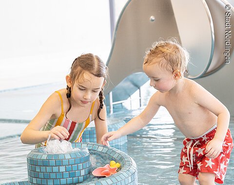 Zwei Kinder beim Spielen im Kinderbereich im Kraterbad Zwei Kinder spielen in einem flachen Schwimmbecken an einem blauen Mosaikbrunnen mit Wasserspielzeug.