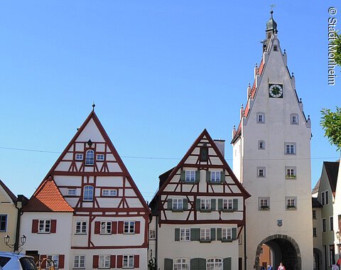 Historische Fachwerkhäuser, ein Torturm mit Uhr in einer Altstadt bei klarem Himmel. Links im Bild parkende Autos auf einem gepflasterten Platz. Rechts im Vordergrund ein Baum mit einer Info-Tafel und einem großen Buchstaben "O" aus Stein. Dahinter eine Informationsvetrine.
