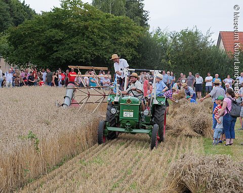 Grüner Traktor mit Erntegerät fährt durch Getreidefeld, viele Menschen beobachten die Ernteaktion.