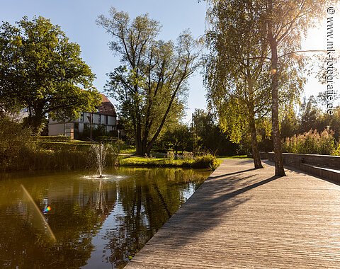 Holzsteg am Weiher mit Springbrunnen, Bäumen und Haus im Hintergrund bei Sonnenschein am Nachmittag