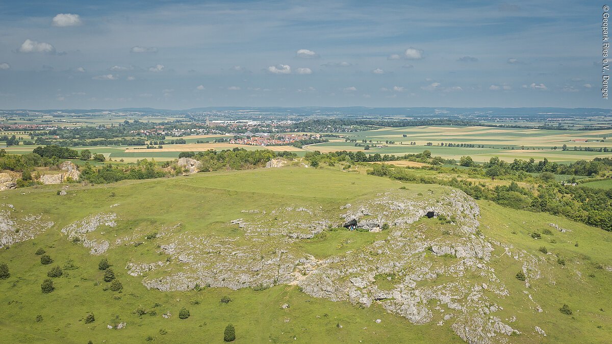 Luftbildaufnahme vom Riegelberg mit den Ofnethöhlen Hügelige Landschaft mit grünen Wiesen und Felsen im Vordergrund, Felder und ein Dorf im Hintergrund.