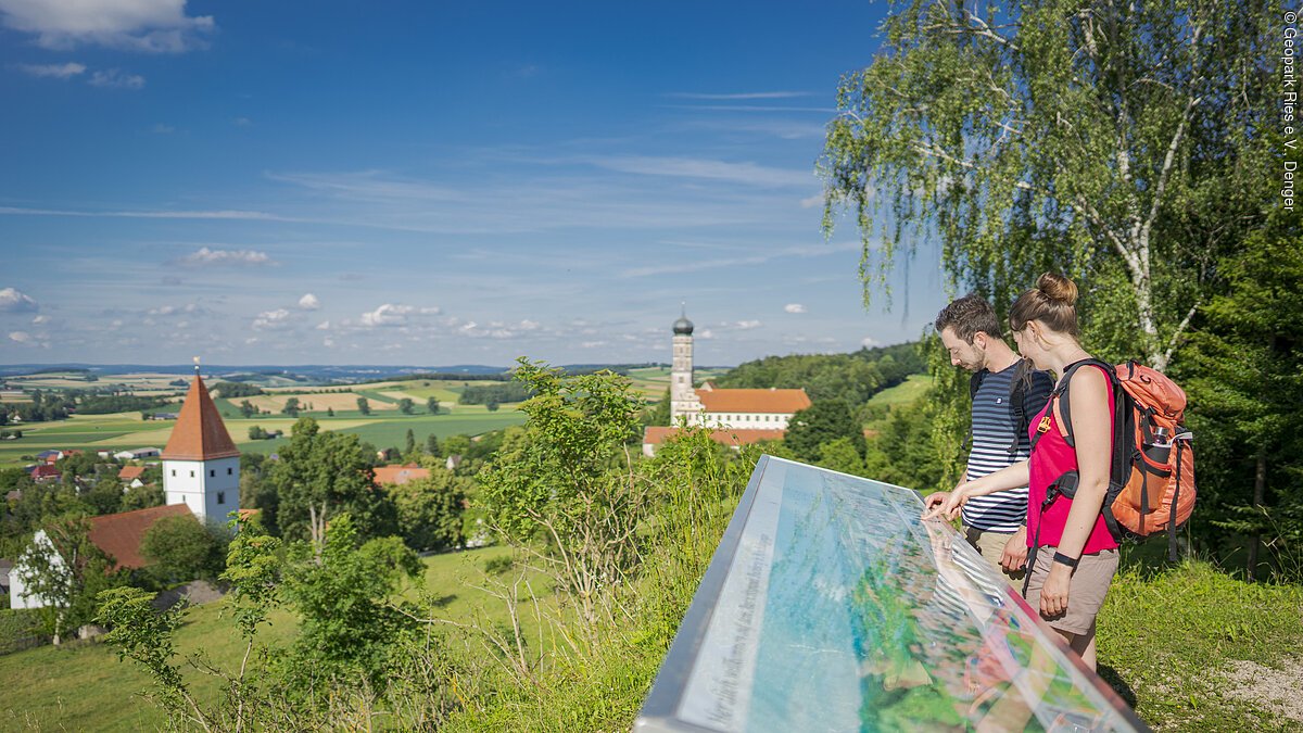 Zwei Personen betrachten eine Informationstafel mit Blick auf eine ländliche Landschaft mit Kirche und Kloster.