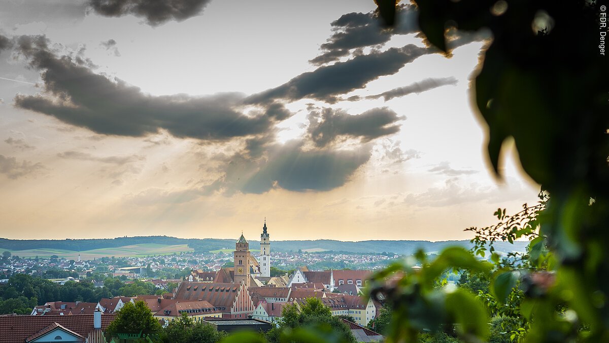 Stadtansicht Donauwörths mit Kirchtürmen der Liebfrauenkirche und des Klosters Heilig Kreuz im Vordergrund, bewölkter Himmel und Sonnenstrahlen im Hintergrund.