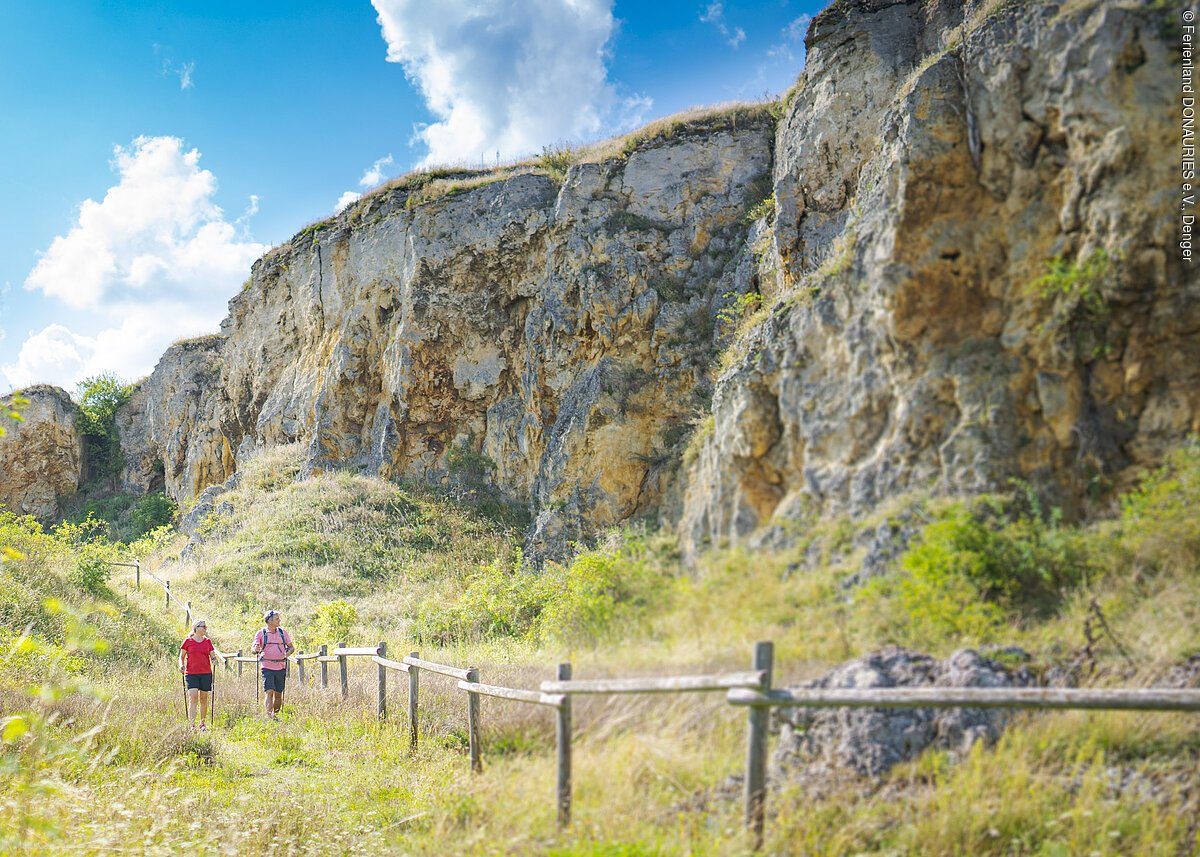 Zwei Personen wandern auf einem Pfad entlang einer felsigen Klippe bei sonnigem Wetter.