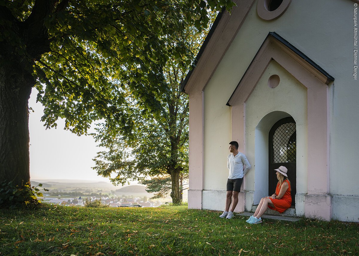 Wanderer auf der Monheimer Alb Mann lehnt an Hauswand, Frau sitzt in Türbogen, große Bäume, Dorf im Hintergrund bei Tageslicht.