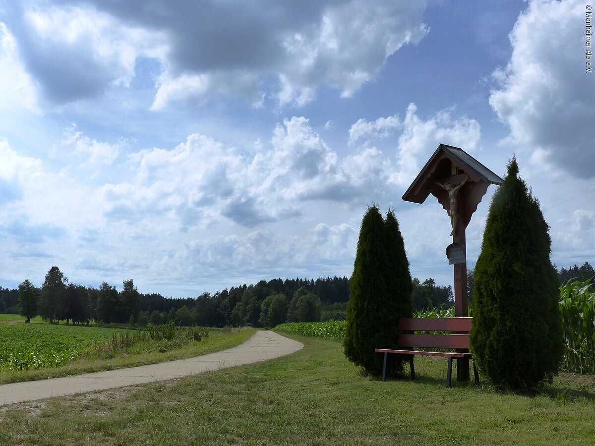 Blick auf ein idyllisches Fleckchen - ein Wegkreuz mit Holzbank davor - im Hintergrund sieht man den Wald.