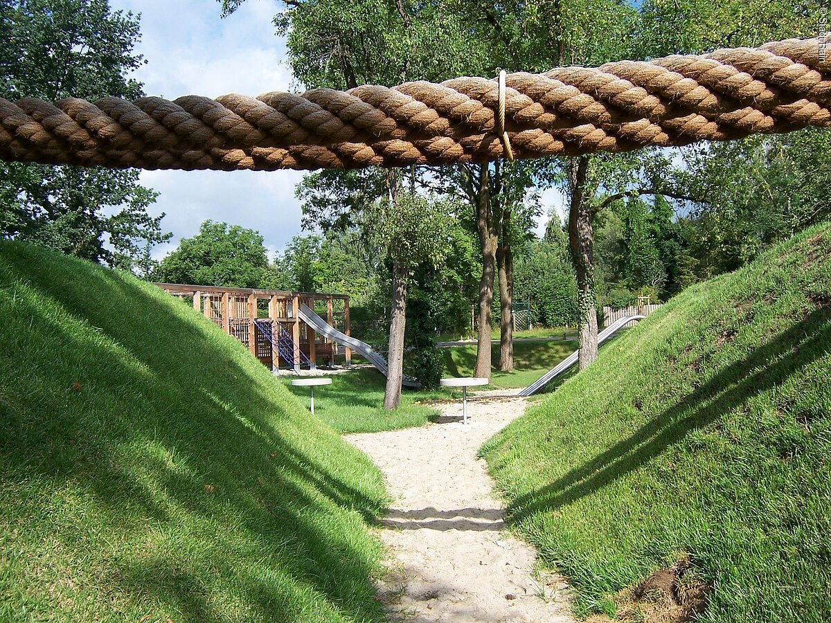 Blick auf die im Grünen, unter Bäumen liegenden Spielgeräte.