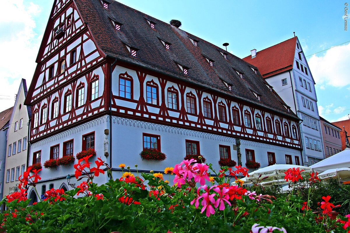 Fachwerkhaus mit roten Balken und Blumenkästen, davor bunte Blumen und Sonnenschirme bei blauem Himmel.