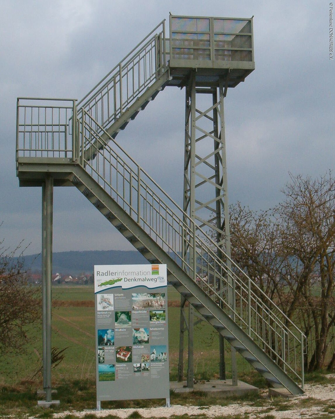 Blick auf den Aussichtsturm am Denkmalweg mit davor stehender Infotafel.