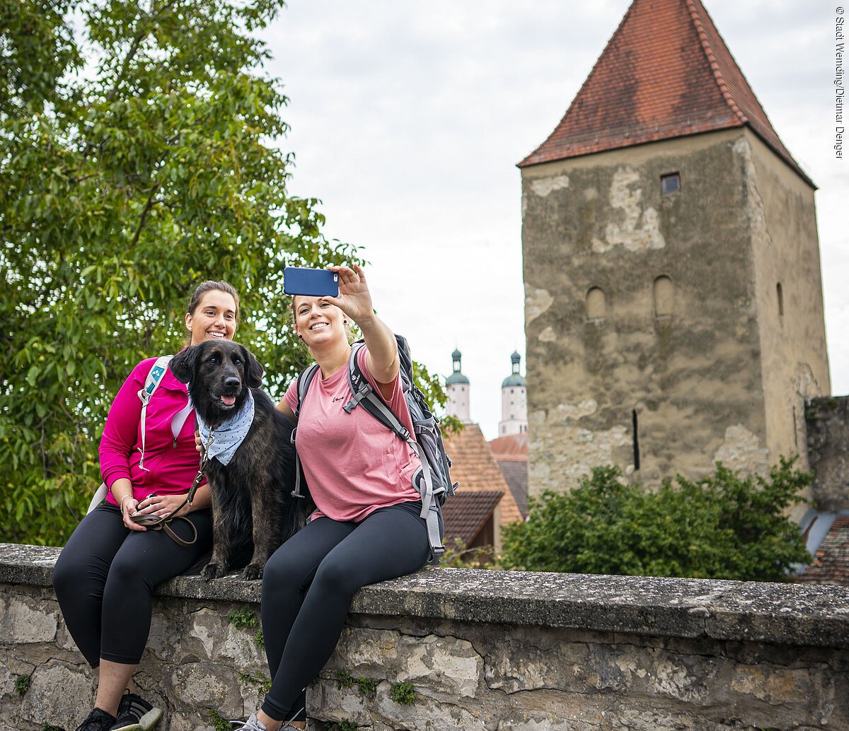 Zwei Freundinnen sitzen mit ihrem Hund auf der Stadtmauer und machen ein Selfie mit dem Baronturm.
