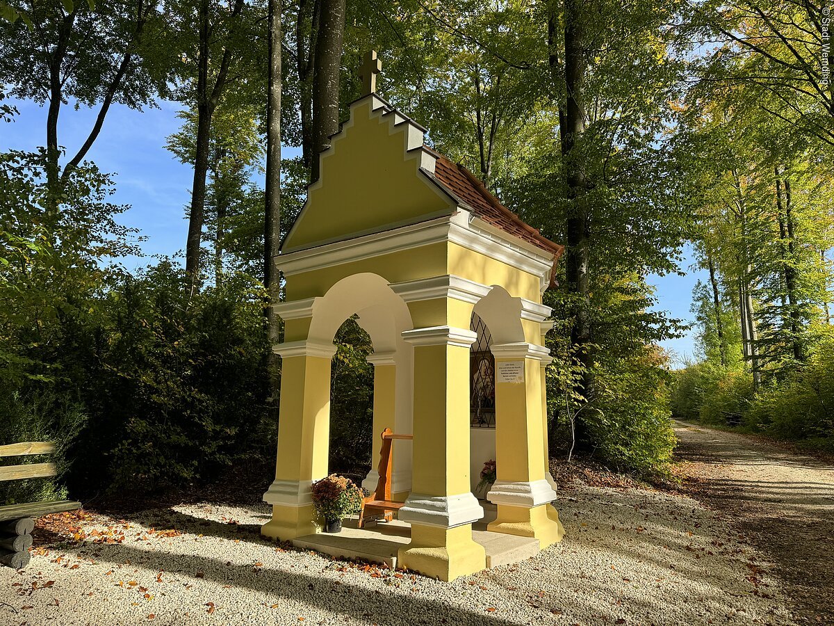 Eine gelbe kleine Kapelle steht an einem Waldweg. Außemherum sind grün belaubte Bäume.