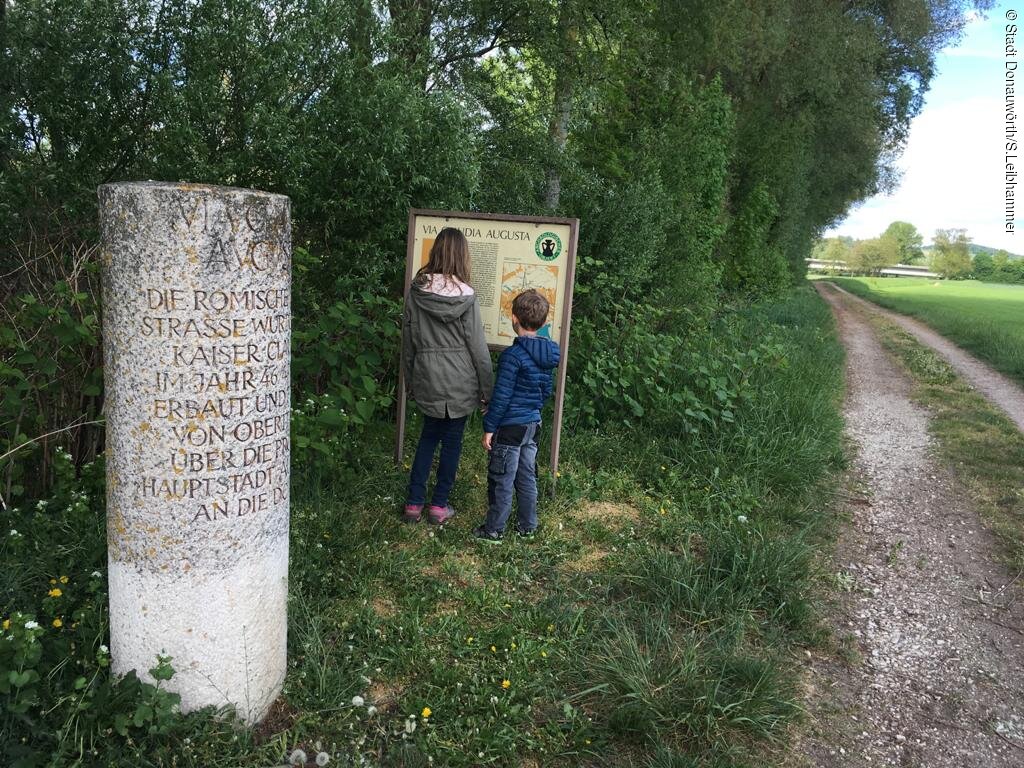 Blick von zwei Kindern auf die Informationstafel und daneben der Meilenstein der Römerstraße Via Claudia Augusta.