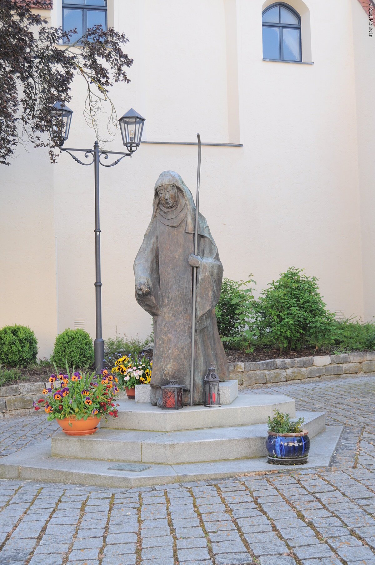 Steinstatue der heiligen Walburga in Monheim mit Stab, umgeben von Blumen in Töpfen und Laternen, vor einer hellen Gebäudewand, der Stadtpfarrkirche Monheim. Vor dem Gebäude sind Pflanzen eingesetzt.