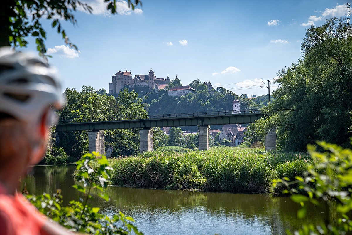 Radfahren an der Wörnitz mit Burgblick Blick auf die Eisenbahnbrücke über der Wörnitz mit der Harburg im Hintergrund, Fahrradhelm einer Radfahrerin im Vordergrund.