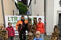 Vier Personen stehen hinter und neben geschnitzten Holzfiguren von Tieren auf einem gepflasterten Platz und blicken in die Kamera. Im Hintergrund zwei Wohnhäuser und eine kleine Gasse zu sehen, sowie ein Werbebanner an einem Bauzaun.