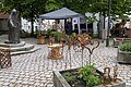 Marktstand mit Metallkunstwerken auf dem Pflasterplatz verteilt, Person am Pavillon mit schwarzem Dach, Marienstatue links, Bäume und Wohnhäuser im Hintergrund.