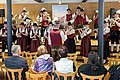 Blaskapelle in rot/schwarz/weißer Tracht spielt auf der Bühne, mit einer Holzverkleidung im Hintergrund, davor sitzen sechs Zuschauer auf Holzstühlen.