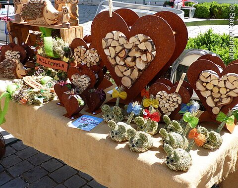 Marktstand mit Holzherzen, Holztieren und dekorativen Figuren aus Stroh auf einem mit Leinen bedeckten Tisch.
