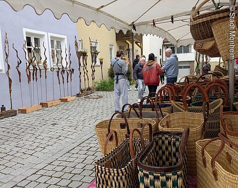 Marktstand in einer Innenstadt mit geflochtenen Körben, im Hintergrund Menschen und dekorative Metallstäbe vor bunten Häuserwänden.