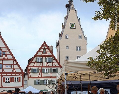 Markt mit Ständen und Besuchern vor Fachwerkhäusern und einem Turm mit Uhr und Storchnest.